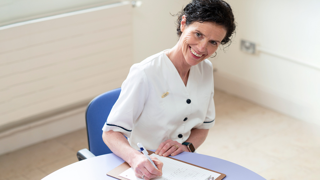 Ann Noonan in uniform sitting at a table with pen and paper looking at camera and smiling